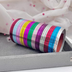 Op de afbeelding: Een stapel kleurrijke armbanden in tinten roze, blauw, groen, geel en zilver. De armbanden zijn in een cilindrische vorm gerangschikt en liggen op een grijze fluwelen tray. Een doorschijnende witte stof met roze stippen is op de achtergrond.