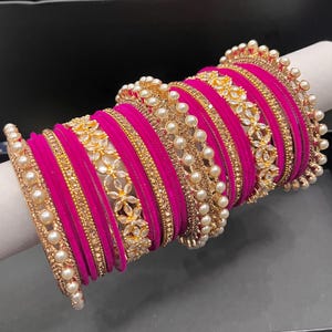May include: A set of fuchsia bangles featuring alternating bands of plain velvet-textured material, gold-toned metal studded with small clear stones, and gold-toned metal accents decorated with white faux pearls and floral-shaped clear stones.