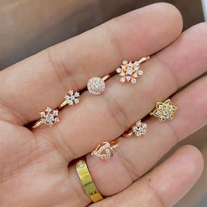 May include: A collection of gold-toned nose rings and studs featuring clear cubic zirconia stones in various shapes, including snowflakes, flowers, a circular cluster, and a triangular knot design, alongside a plain gold band ring.