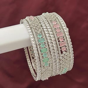 Pode incluir: Uma pilha de pulseiras de prata com pedras preciosas claras e coloridas. As pulseiras são adornadas com pequenas pedras claras e pedras retangulares rosa e verdes maiores. As joias são exibidas em um fundo vermelho escuro.
