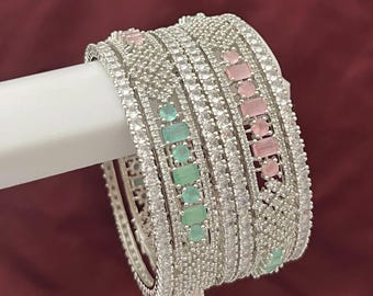 May include: A stack of silver bangles with clear and colored gemstones. The bangles are embellished with small, clear stones and larger, rectangular pink and green gems. The jewelry is displayed against a dark red background.