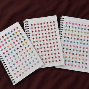 May include: Three spiral-bound notebooks with white pages featuring grids of small, circular, multi-colored stickers. The stickers are arranged in neat rows and columns, displaying a variety of colors including red, blue, green, yellow, and pink.