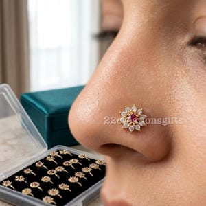 Puede incluir: Un piercing de nariz dorado con un diseño floral, con una piedra roja central rodeada de gemas blancas. La imagen también muestra una colección de piercings de nariz similares en una caja de plástico transparente.