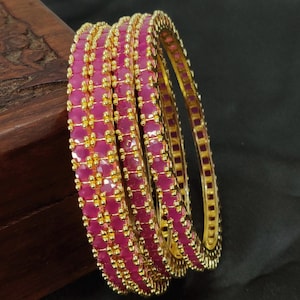 May include: A set of three gold-toned bangles featuring rows of faceted, vibrant pink stones. Each bangle is constructed with a metallic frame that holds the stones in place, accented by small, raised gold-coloured beads between the pink segments.