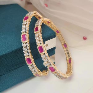 May include: Two gold bangles with alternating rectangular pink gemstones and small clear stones. The bangles are displayed on a teal surface with a white and pink background. The jewelry is designed with a classic, elegant style.