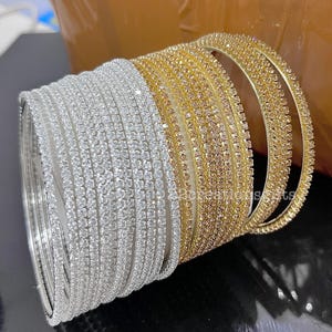 Può includere: Una pila di braccialetti argentati e dorati. I braccialetti argentati sono a sinistra e quelli dorati a destra. Ogni braccialetto è incastonato con piccole pietre scintillanti. I braccialetti sono impilati.