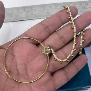 Puede incluir: Un aro nasal dorado con un diseño circular, un adorno central en forma de flor con piedras transparentes y una cadena decorada con pequeñas cuentas blancas y esferas doradas, medido con una regla que muestra aproximadamente 5 cm.