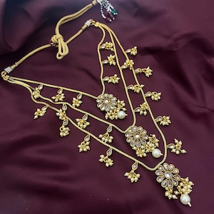 Puede incluir: Collar dorado con múltiples hilos y colgantes florales. El collar presenta pequeñas perlas blancas y gotas de perlas más grandes. El diseño incluye un cordón decorativo y está sobre un fondo burdeos oscuro.