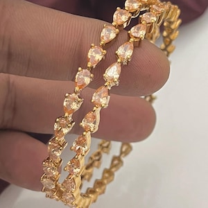 Può includere: Due braccialetti color oro con un motivo ripetuto di pietre preziose a forma di goccia di colore pesca. I braccialetti sono presentati su uno sfondo neutro, evidenziando il design e il colore dei gioielli.