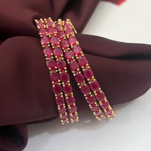 May include: Four gold bangles, each adorned with oval, ruby-red gemstones. The bangles are arranged to overlap slightly, highlighting the vibrant colour of the stones. The gold setting provides a warm contrast to the deep red hues of the gemstones.