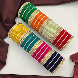 Può includere: Due pile di braccialetti colorati. Una pila presenta un gradiente di verde, arancione e verde chiaro. L'altra pila ha un gradiente di giallo, turchese, rosa e blu navy. Ogni braccialetto è adornato con abbellimenti color oro.