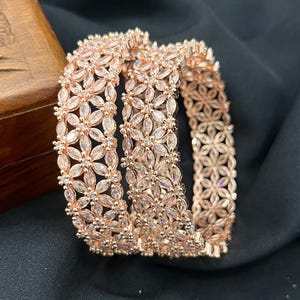 Peut inclure: Deux bracelets joncs en or rose arborant un motif floral répétitif composé de pierres claires taillées en marquise et rondes. La structure métallique est délicate et ajourée, présentant un agencement continu de fleurs serties de cristaux blancs.