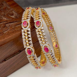 May include: Two gold bangles featuring clear and red gemstones. The bangles have an intricate design with oval red stones and smaller clear stones. The jewellery is displayed on a wooden surface.