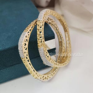May include: Two gold bangles with detailed openwork patterns. Each bangle has a row of small, sparkling stones. The bangles are displayed on a teal surface, with a soft, white background.