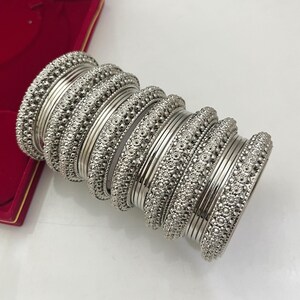 Pode incluir: Um conjunto de pulseiras prateadas dispostas em uma pilha. A coleção apresenta designs alternados, incluindo faixas lisas e polidas e faixas texturizadas com um padrão circular repetitivo. O acabamento metálico reflete a luz na superfície.