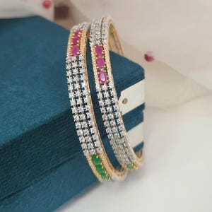 Op de afbeelding: Twee gouden armbanden met een rij heldere stenen en afwisselend rode en groene stenen. De armbanden worden gepresenteerd op een turquoise oppervlak met een witte achtergrond.