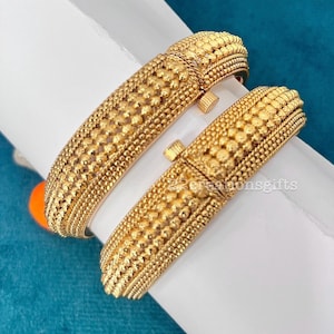 May include: Two gold-toned bangles featuring a textured surface with rows of small raised beads and a central cylindrical clasp mechanism. The metal has a matte finish with intricate detailing across the entire exterior of each circular bracelet.