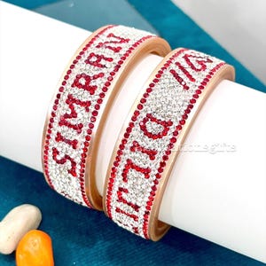 Puede incluir: Dos brazaletes metálicos con una banda ancha incrustada con pedrería blanca transparente y roja que forma los nombres Simran y Diljit. Las pulseras tienen un acabado liso y pulido en los bordes y una estructura rígida.