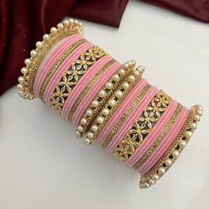 May include: A collection of pink bangles with gold and pearl detailing. The bangles have a velvety texture and are decorated with gold floral patterns and rows of small pearls. The edges of the bangles are also lined with pearls.