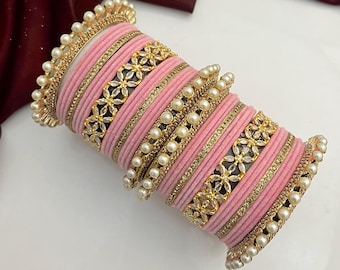 May include: A stack of pink bangles with gold and pearl accents. The bangles have a velvet-like texture and are adorned with gold floral designs and rows of small pearls. The edges of the bangles are also lined with pearls.