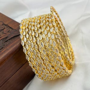 Puede incluir: Un conjunto de pulseras doradas, cada una adornada con numerosas piedras claras de forma ovalada. Las pulseras están dispuestas en una formación curva, mostrando su intrincado diseño y su apariencia brillante. Las joyas están sobre una superficie de madera.