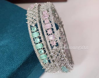 May include: Two silver bangles with alternating pastel pink and mint green rectangular stones. The bangles are encrusted with small, clear crystals. The jewelry is displayed on a teal surface.