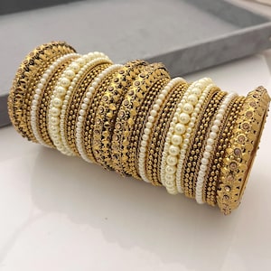 May include: A collection of gold bangles with detailed patterns and embellishments. Some bangles feature rows of small pearls, while others have detailed floral designs. The bangles are stacked together, creating a rich and ornate appearance.