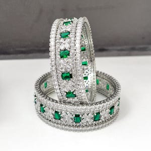 Pode incluir: Duas pulseiras de prata com um design brilhante. Cada pulseira apresenta uma fileira de pedras retangulares verde-esmeralda intercaladas com pedras claras menores. As pulseiras estão sobre uma superfície branca com fundo cinza.
