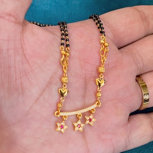 Puede incluir: Collar dorado con cadena de cuentas negras. El colgante presenta una barra curva con pequeñas piedras transparentes y tres colgantes en forma de estrella con centros rojos. También hay colgantes dorados en forma de corazón y redondos.