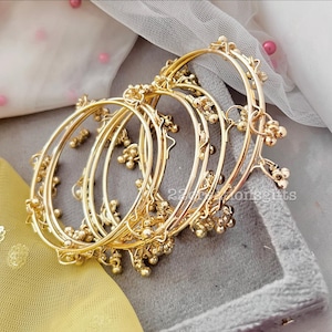 May include: A collection of gold-colored bangles with small, dangling bell-shaped ornaments. The bangles are arranged in a stack, showcasing their intricate design and reflective surface. The background includes soft, draped fabric.