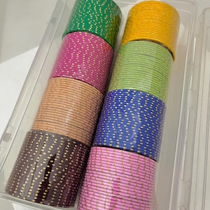 Puede incluir: Un recipiente de plástico transparente contiene pilas de pulseras coloridas. Las pulseras son en tonos de verde, rosa, melocotón, marrón, amarillo, verde claro, azul y rosa. Cada pulsera está decorada con un patrón de puntos dorados.