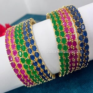 Puede incluir: Un conjunto de brazaletes dorados con filas de piedras preciosas de corte ovalado en tonos vibrantes de rosa, verde y azul. Cada brazalete está engastado con piedras, creando un patrón colorido continuo. El texto 22creationsgifts es visible.