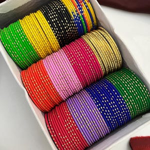 May include: A box filled with colorful bangles. The bangles are in various colors, including green, yellow, black, gold, pink, red, purple, blue, and gold. Each bangle is adorned with small, gold-colored dots. The bangles are neatly arranged in rows within the box.