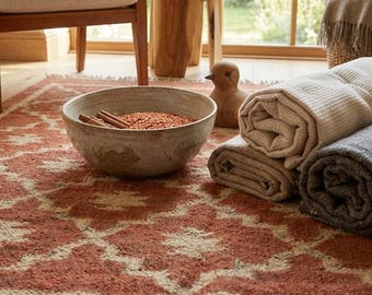 Puede incluir: Una alfombra con estampado en terracota y crema con bordes de flecos, que incluye un cuenco de cerámica grande lleno de lentejas rojas y ramas de canela, una figura de pájaro de madera y dos mantas texturizadas enrolladas.