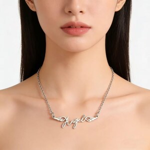 May include: Silver-toned necklace featuring the word "Angel" in a cursive script, with small wing designs on either side. The chain is a delicate link style, and the necklace is worn around the neck.