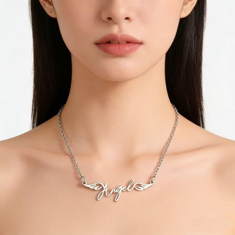 May include: Silver-toned necklace featuring the word "Angel" in a cursive script, with small wing designs on either side. The chain is a delicate link style, and the necklace is worn around the neck.