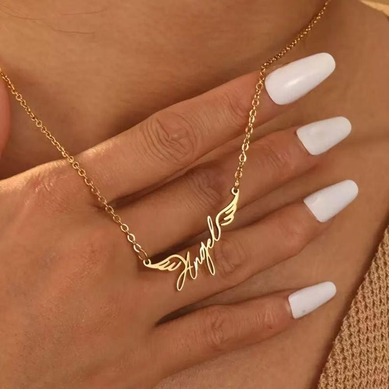 May include: A gold-colored necklace with the word "Angel" in cursive script, flanked by small wing designs. The necklace is on a delicate chain. The image shows the necklace on a person's hand.