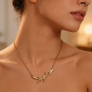 May include: A gold-colored necklace with the word "Angel" in cursive script and small wing designs on either side. The necklace is worn around the neck and is made of a delicate chain.