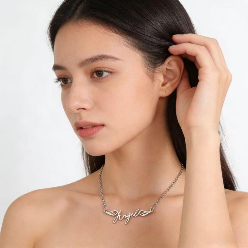 May include: A silver-colored necklace with the word "Angel" in cursive script and small wing designs on each side. The necklace is on a delicate chain.