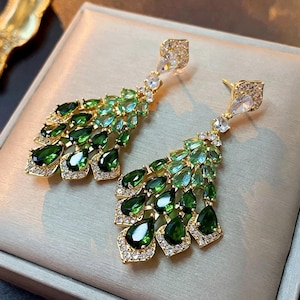 Puede incluir: Un par de pendientes dorados con un diseño en cascada. Los pendientes presentan una mezcla de piedras preciosas en forma de lágrima verde esmeralda y verde claro, acentuadas con cristales transparentes. Cierre de poste.