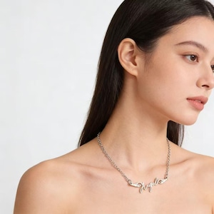 May include: Silver-toned necklace with the word "Angel" in cursive script, flanked by small wing designs. The necklace is on a chain and is worn around the neck. The background is a plain white.