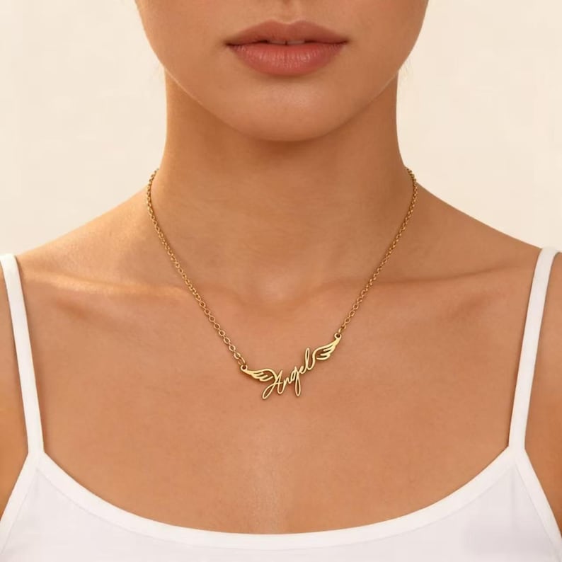 May include: Gold-colored necklace with the word "Angel" in cursive script, flanked by small wing designs. The necklace is worn against a white top.