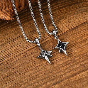 May include: Two silver-toned necklaces with star-shaped pendants. One pendant has a black center, while the other has a cross-like design. The necklaces are displayed on a wooden surface, showcasing the jewelry's details.