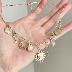 Peut inclure: Collier doré à breloques avec divers pendentifs, dont un coquillage, une clé, un croissant de lune, un soleil, un cœur et un pendentif rond. Le collier est tenu dans une main. Les breloques sont dorées.