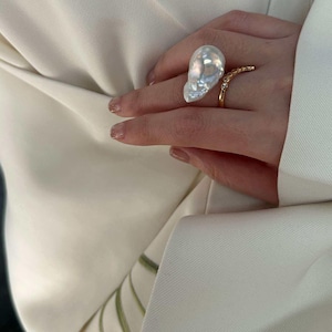 Può includere: Un anello di perla grande, dalla forma irregolare, è il punto focale di questa immagine. La perla ha una lucentezza iridescente. È visibile anche un anello d'oro più piccolo con una fila di piccole pietre. Lo sfondo è un blazer bianco.