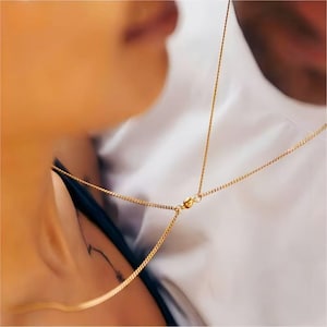 Könnte beinhalten: Eine goldene Körperkette mit filigranem Design. Die Halskette hat mehrere Stränge, die über Schultern und Brust verlaufen. Der Schmuck wird über einem dunklen Oberteil und vor einem weißen Hemd getragen.