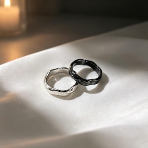 Op de afbeelding: Twee ringen, één zilver en één zwart, met een golvend, organisch ontwerp. De ringen worden gepresenteerd op een witte, zijdeachtige ondergrond. De zwarte ring heeft een glanzende afwerking, terwijl de zilveren ring een metallic glans heeft. De ringen zijn waarschijnlijk van metaal.