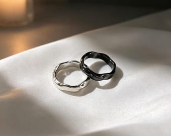 May include: Two rings, one silver and one black, with a wavy, organic design. The rings are displayed on a white, silky surface. The black ring has a glossy finish, while the silver ring has a metallic sheen. The rings are likely made of metal.