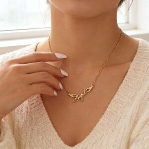 May include: A gold-colored necklace with the word "Angel" in cursive script, flanked by small wing designs. The necklace is worn around the neck, and the chain is visible. The image is a close-up shot.