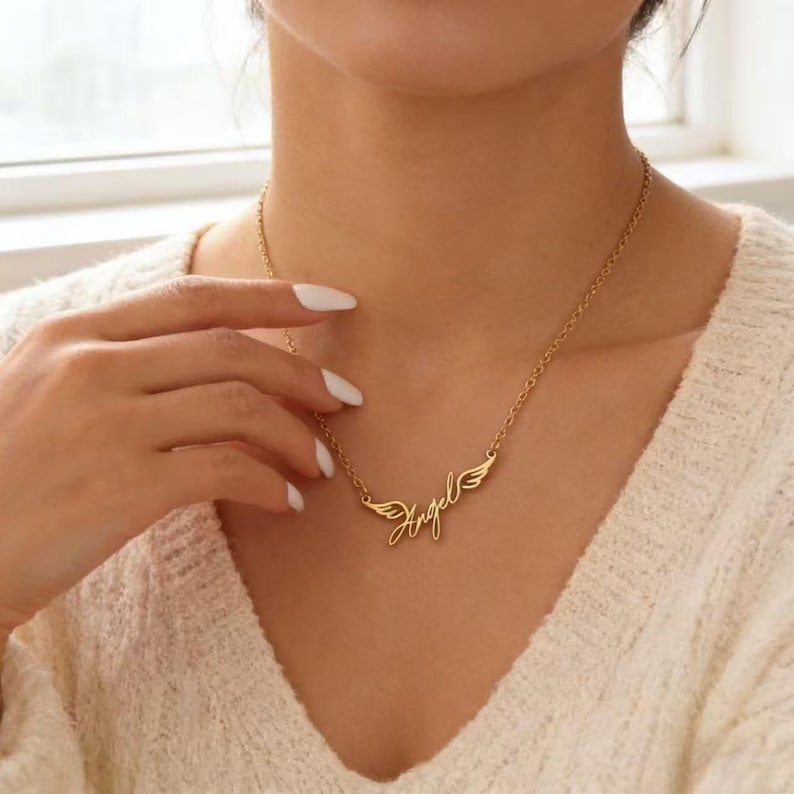 May include: A gold-colored necklace with the word "Angel" in cursive script, flanked by small wing designs. The necklace is worn around the neck, and the chain is visible. The image is a close-up shot.